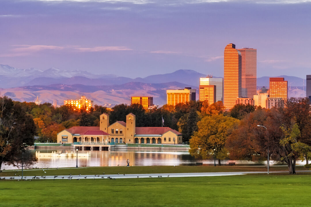 Denver skyline at City Park with water in foreground, skyline mid photo and mountains and sunset in the background